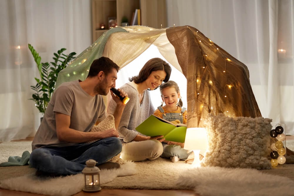 Family sits in a blanket fort with twinkle lights, pillows and lamps. They are reading a book with a flashlight.