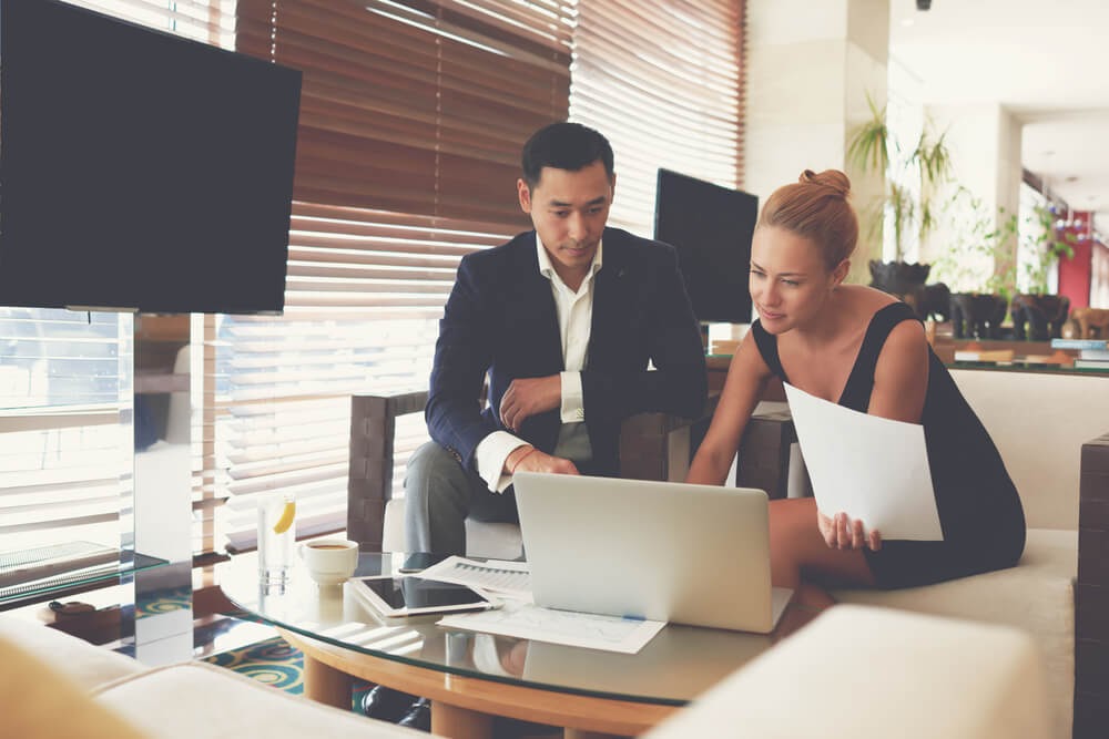 Man and woman sit looking at a laptop and paperwork. Both are dressed professionally.