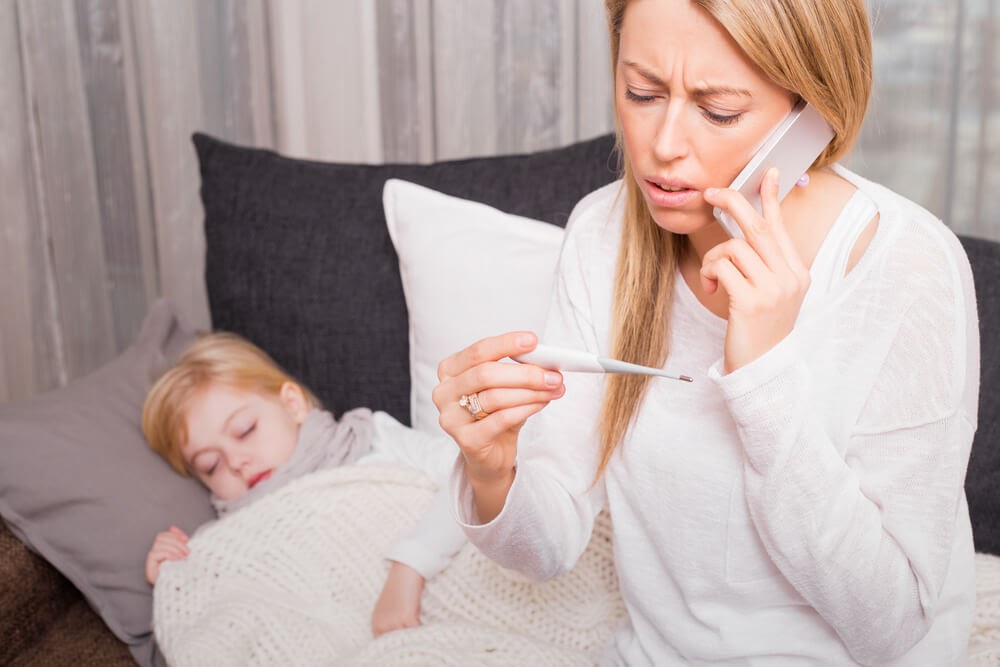 Woman looks at thermometer with worry while talking on the phone. A sleeping child lays on the couch behind her.