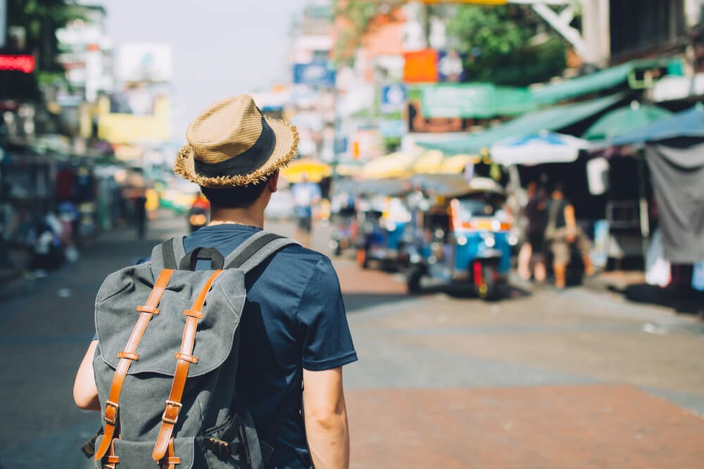 Man standing in a crowded street wearing a straw hat and backpack.