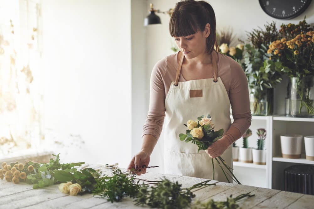Woman arranging flowers wearing a heavy white apron. She is standing in a sun filled room.