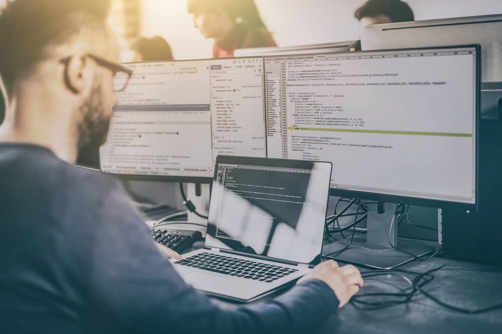Photo of man working on two large computer screens and a laptop