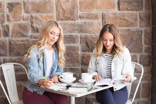 Two women sit at a table with a mugs and books.