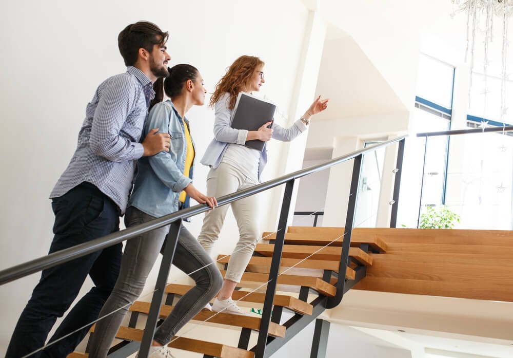 Three people stand on a staircase. One is pointing up the stairs and holds a binder and paperwork.
