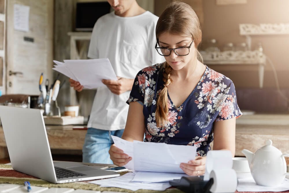 A woman in floral shirt does paperwork and works and a laptop.