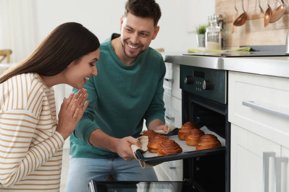Man pulling cinnamon rolls out of the oven and a very happy woman looks on.
