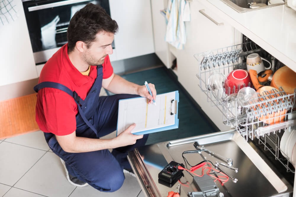 Technician writing up paperwork while looking into a dishwasher.