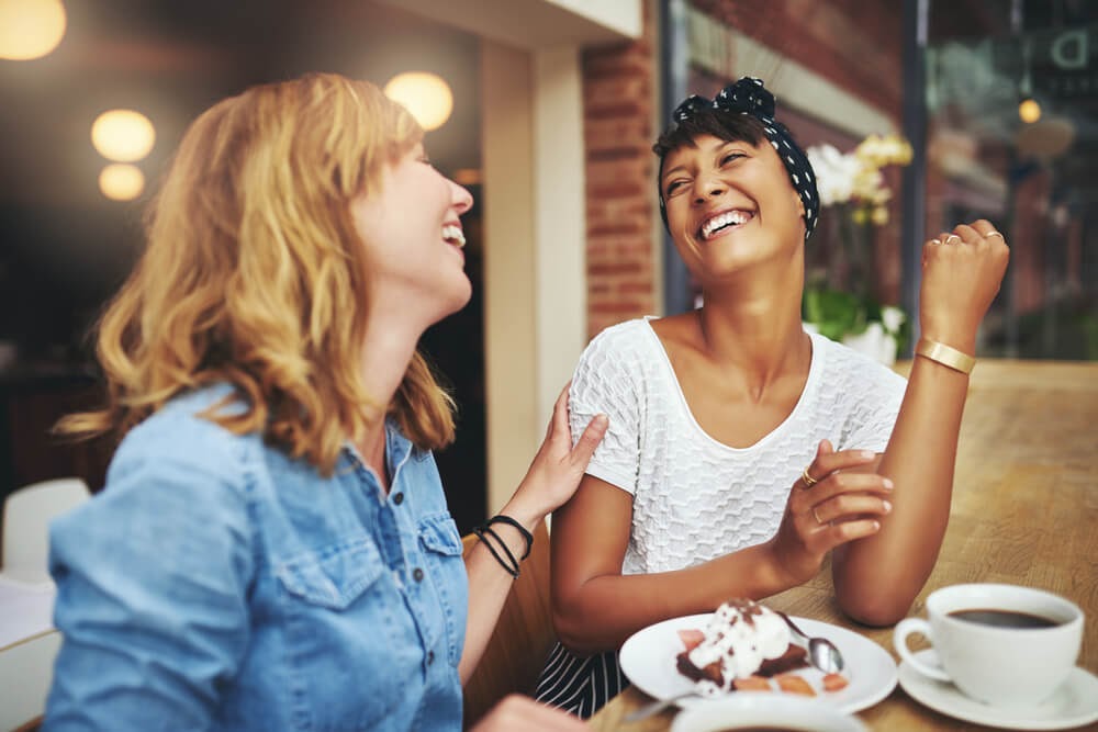 Two women sitting at a table laughing. One wears their hair covered.