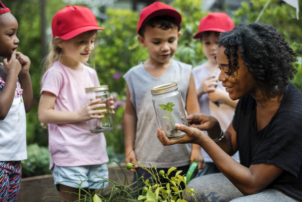 Kids gather around a woman holding a jar and showing them a small plant.