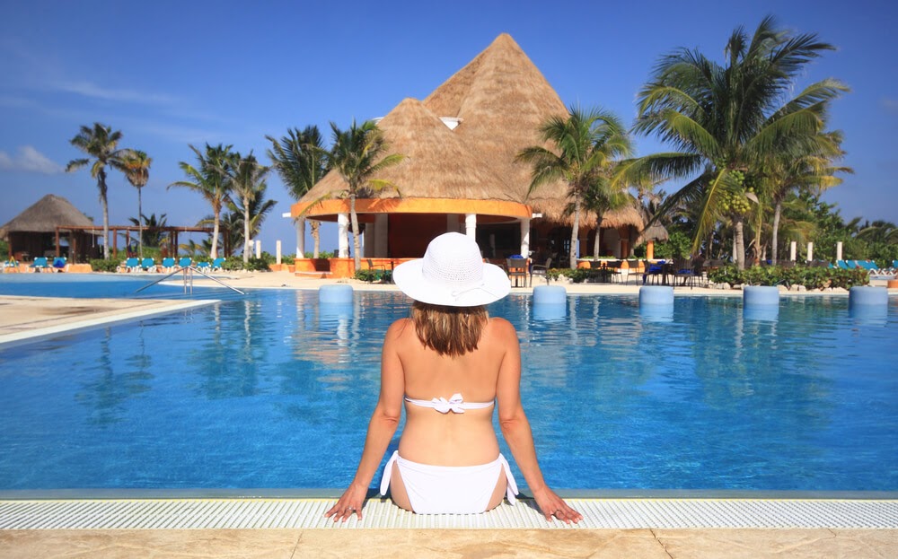 Woman in a white bikini shown sitting poolside from behind.