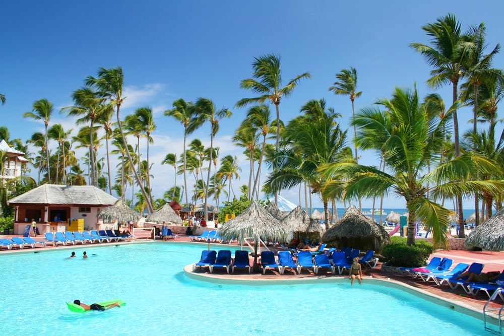 Resort pool with grass cabanas, surrounded by blue lounge chairs.