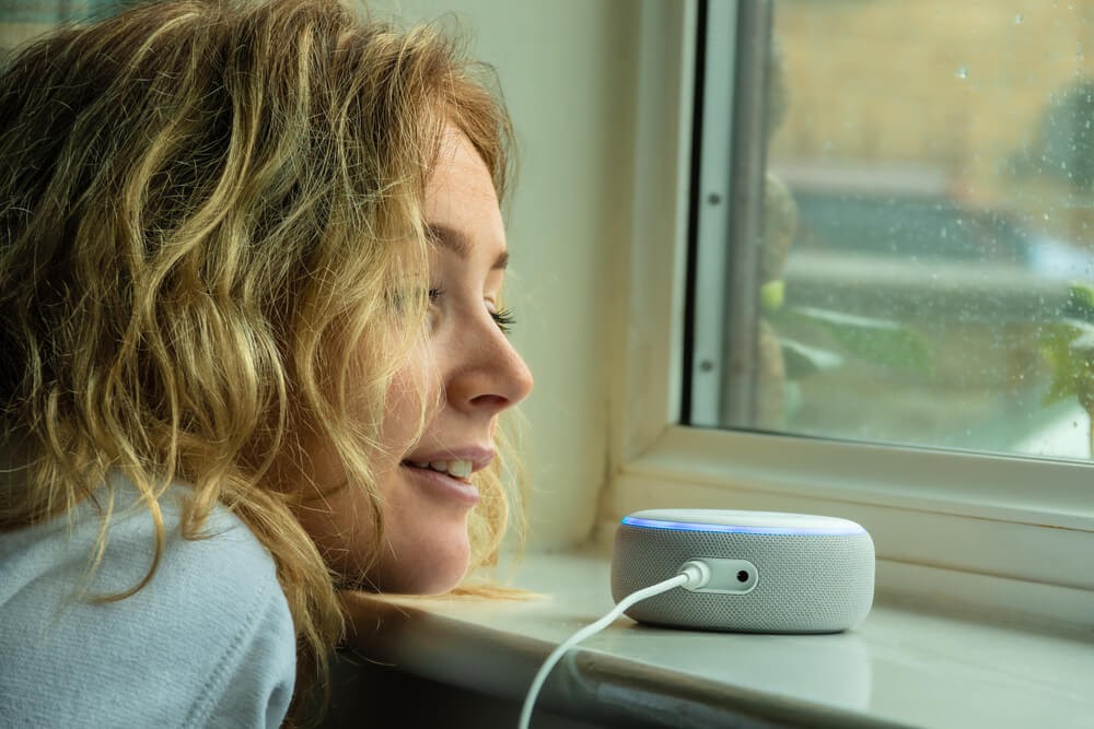 Woman looking out a window with an Alexa speaker near her.