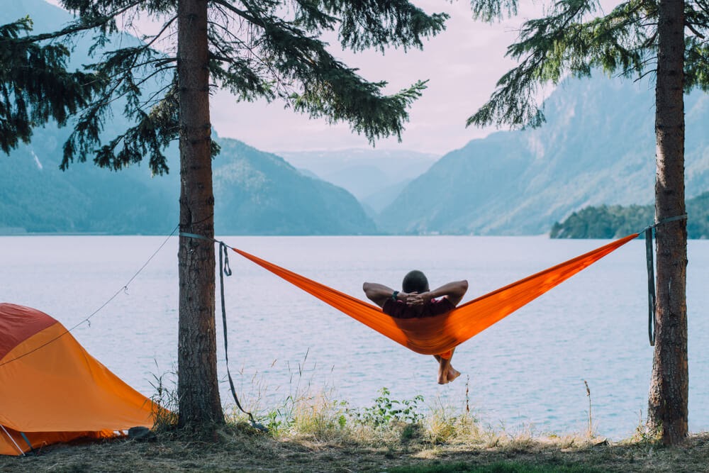 Person lounges in a hammock suspended by two trees and looks out over a lake.