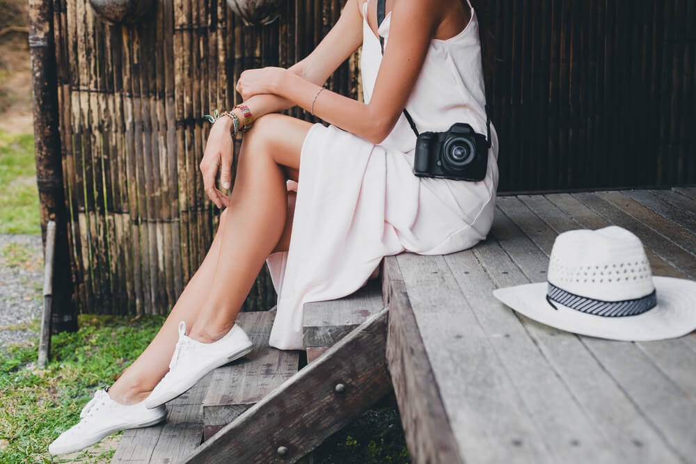 Woman sitting in casual white dress wearing a camera and sneakers.
