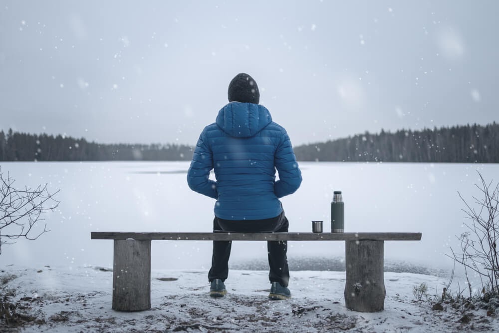 Person from behind sitting on a bench in the snow with a bottle next to them.