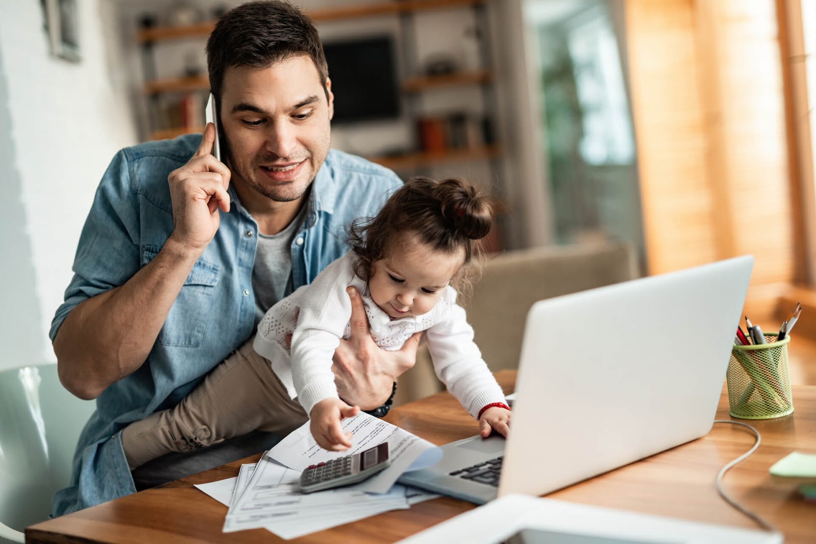 Dad talking on his cell phone while he holds his toddler and tries to work