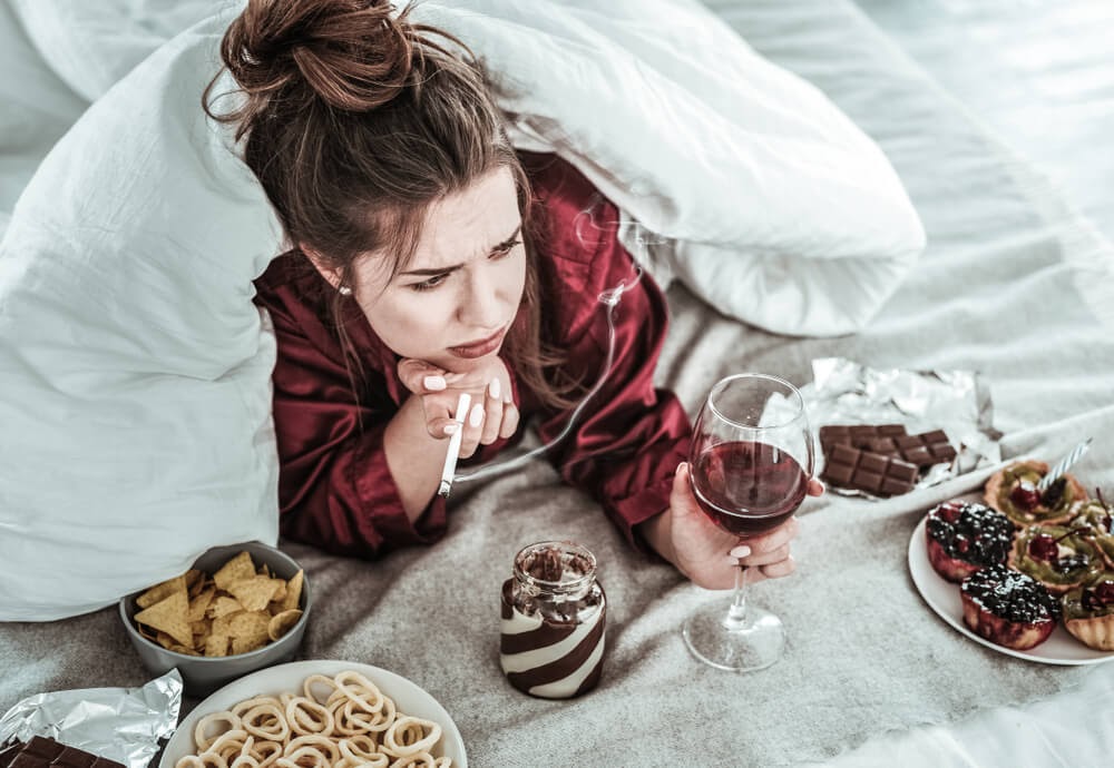 Woman smoking, drinking wine, and eating junk food in bed