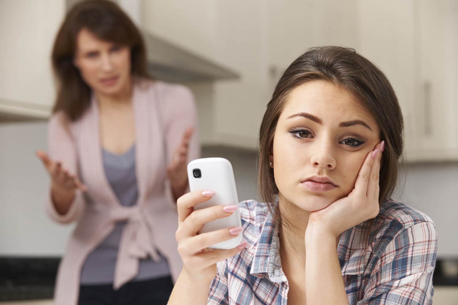 Upset mom in background while teen looks at the camera with a bored expression and phone in here hand