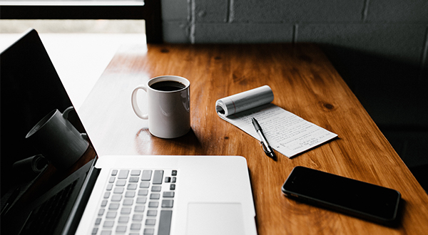 Laptop, coffee cup, cellphone, notepad and pen on a wooden desk