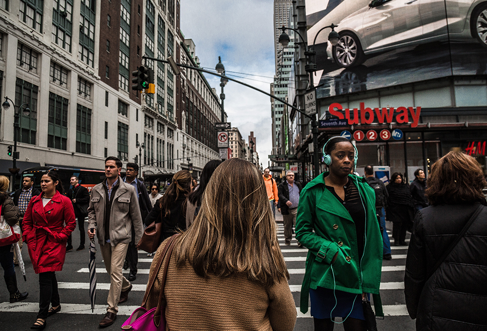 Ethnically diverse pedestrians crossing a street at a crosswalk with a subway station in the background