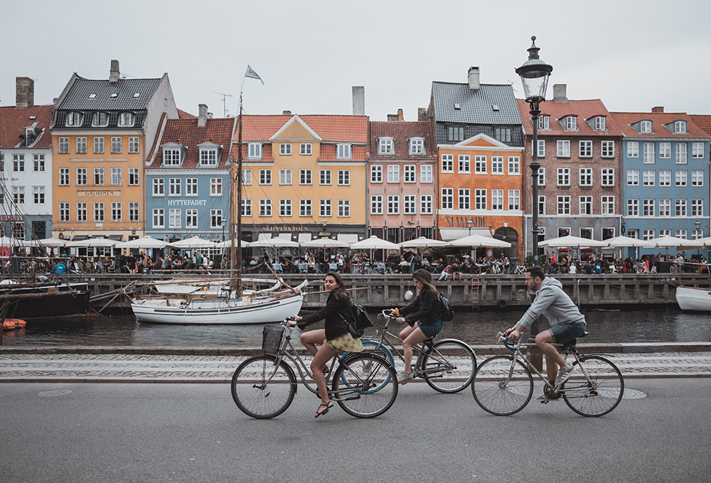 Three people ride bikes with a canal and buildings in the background