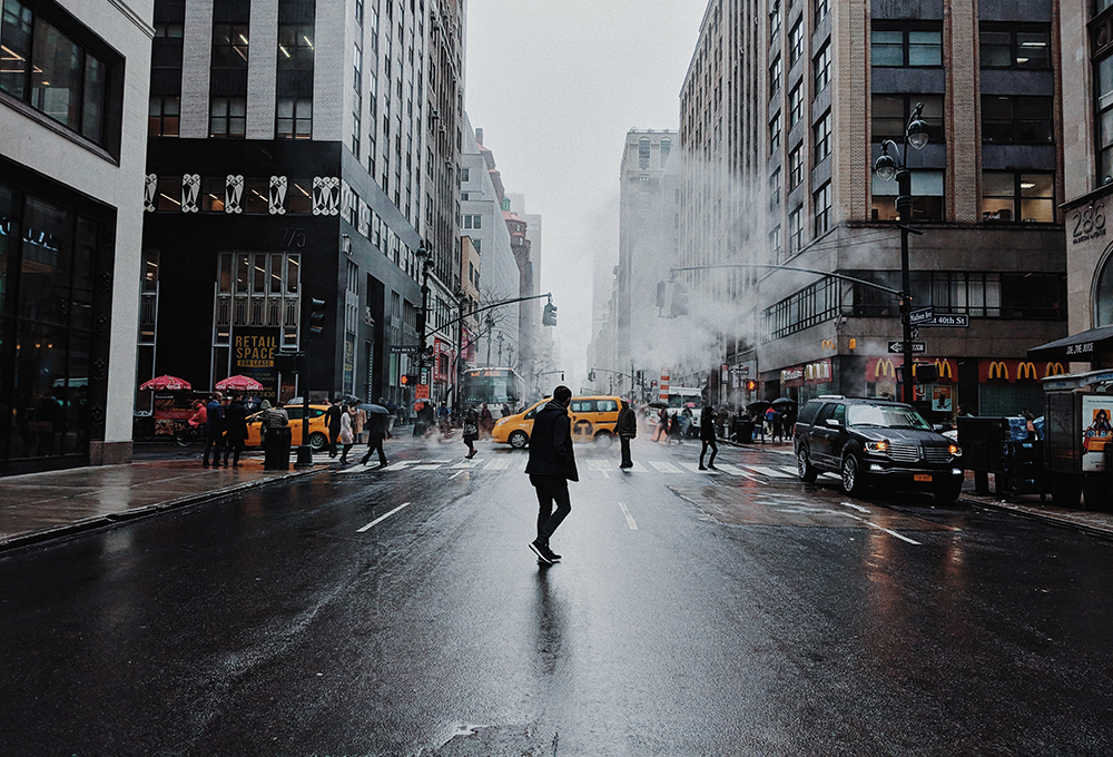 Man crosses wet city street in front of crosswalk with cabs and pedestrians in the backround