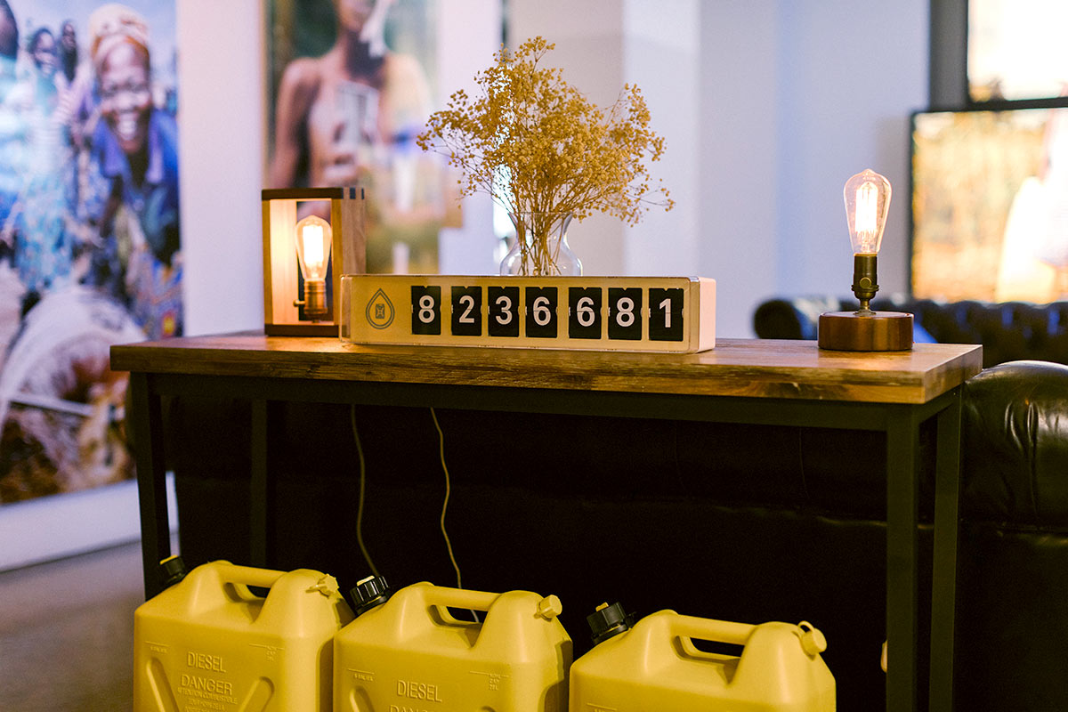 A photo from charity: water's office. A dark brown desk sits in the foreground with three yellow water jugs in front of it. On the desk is a yellow flip counter showing how many wells charity: water has built.