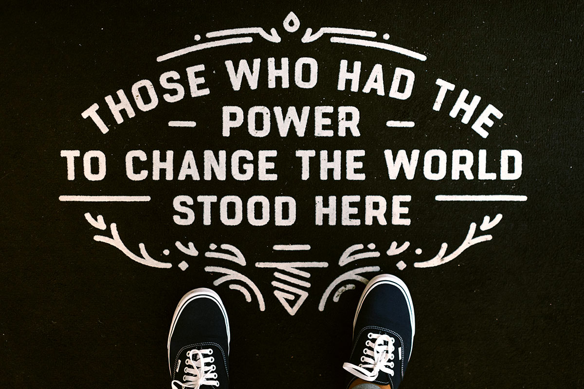 Black background with white script lettering saying, "Those who had the power to change the world stood here." There are black sneakers with white laces at the bottom of the image, making it look like the picture was taken from above.