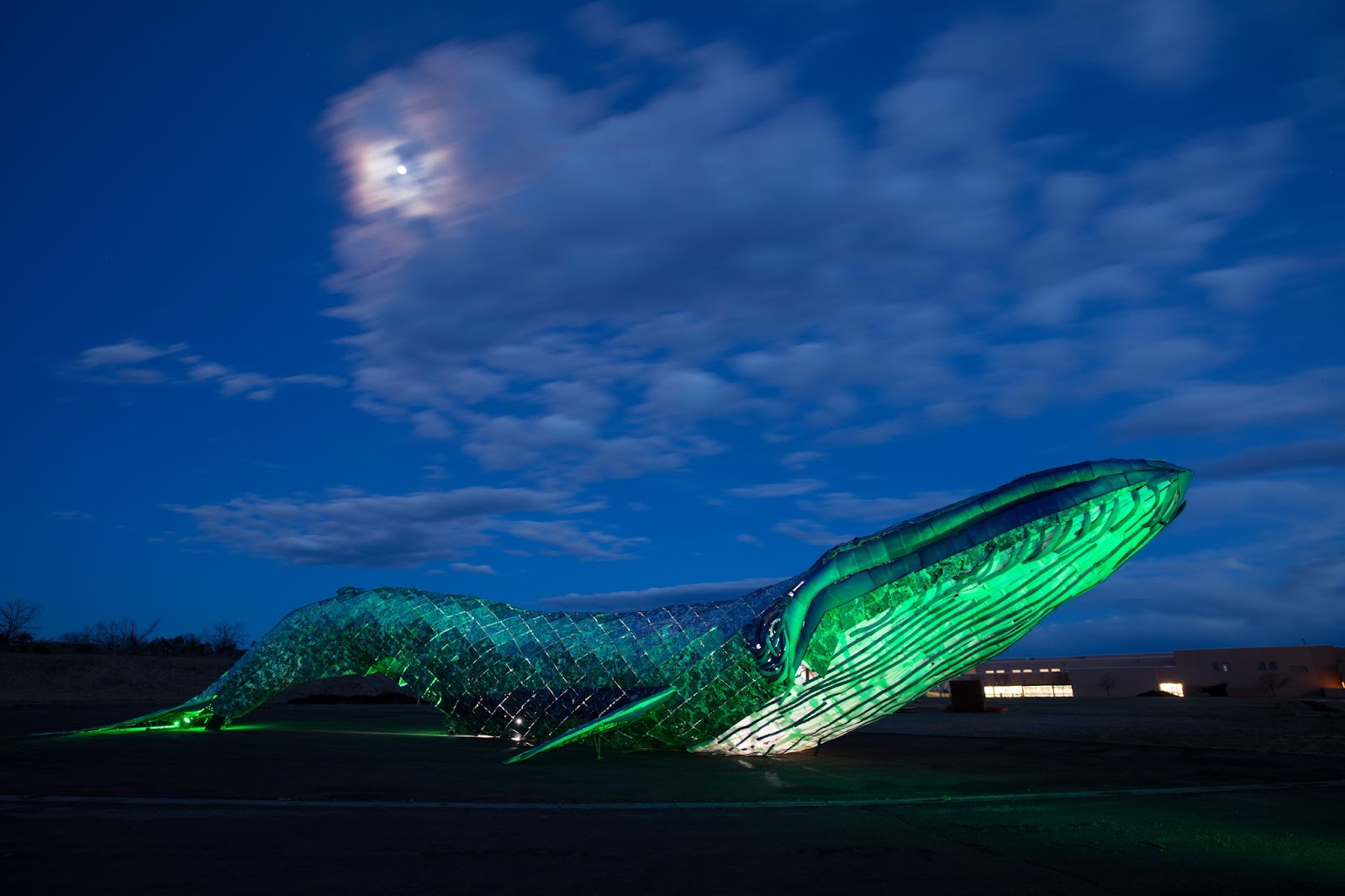 large whale sculpture made of recycled materials with a dark sky and clouds in the background 