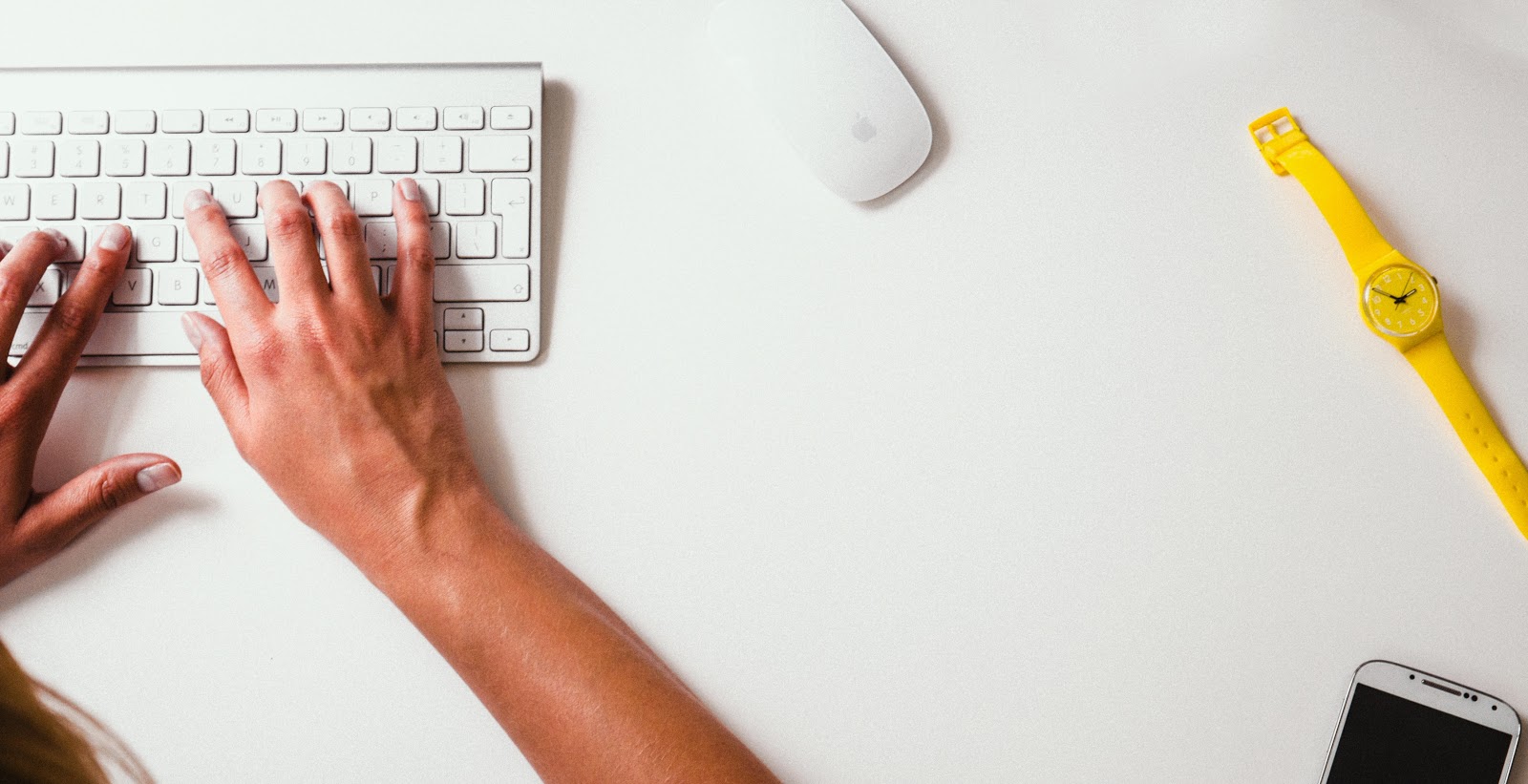 Hands typing at wireless keyboard with mouse, wristwatch, and smartphone on desk