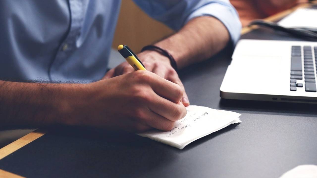 Person writing on a paper, next to a laptop