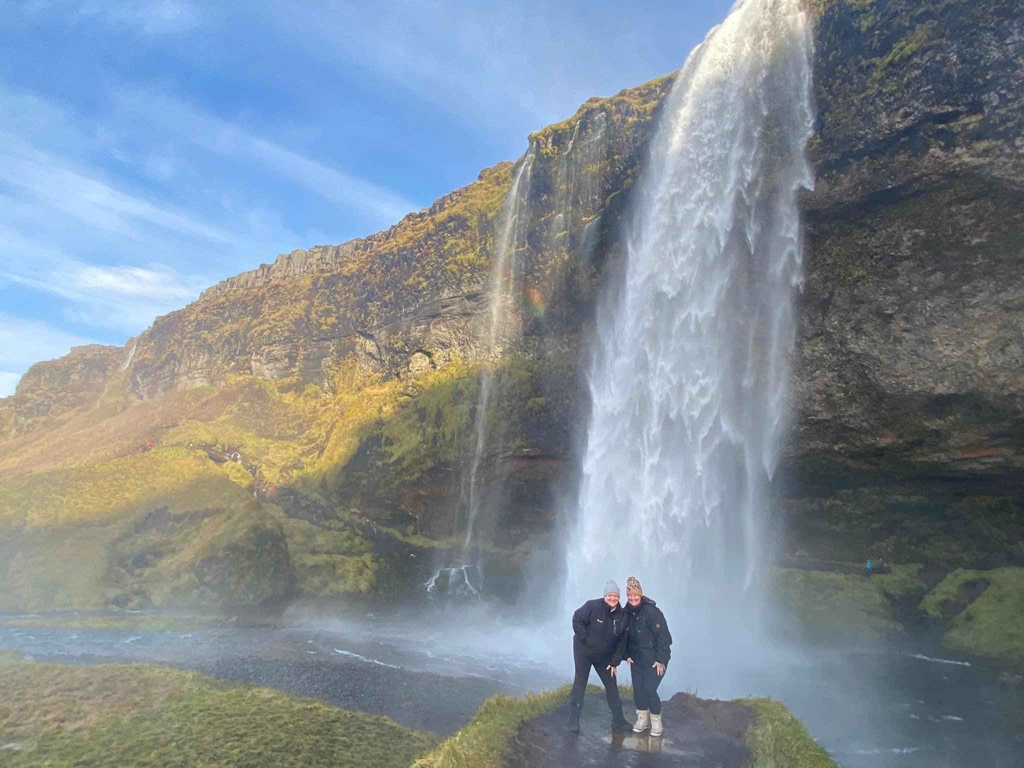 Two people stand on a rock with a waterfall flowing behind them in Iceland.