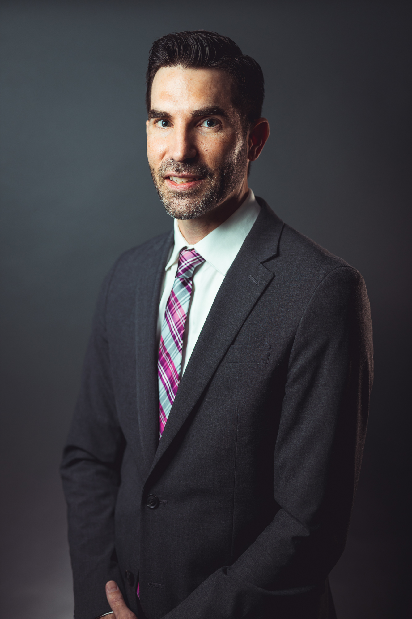 Professional headshot of Erik Merker, Sales Engineer at GRP|WEGMAN, smiling and wearing a dark gray suit coat with a white shirt and colorful plaid tie, in front of a neutral gray background