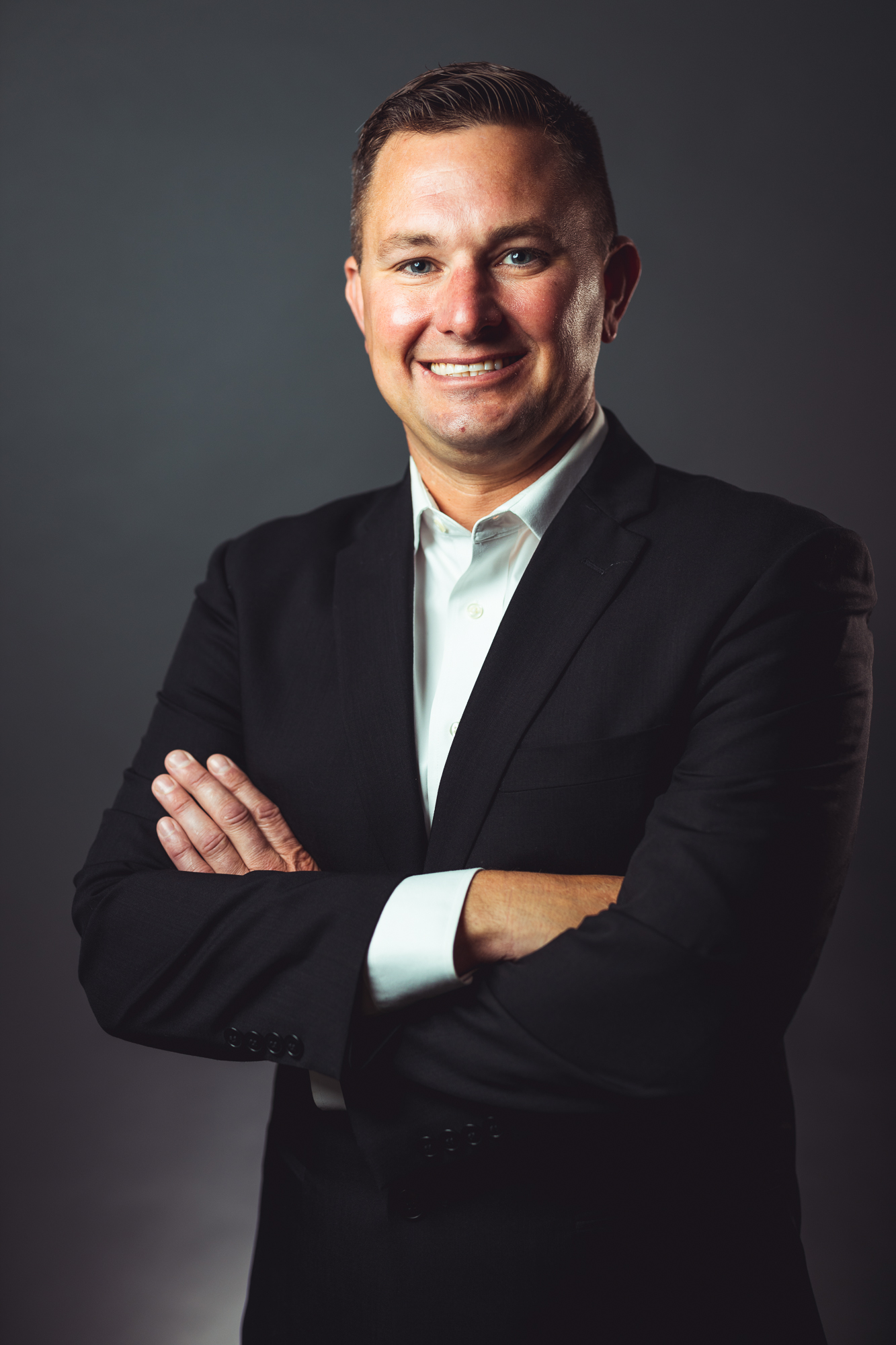 Professional headshot of Chris Fulcher, Director of HVAC Service at GRP|WEGMAN, smiling, arms crossed and wearing a black suit jacket and unbuttoned white shirt, in front of a neutral gray background