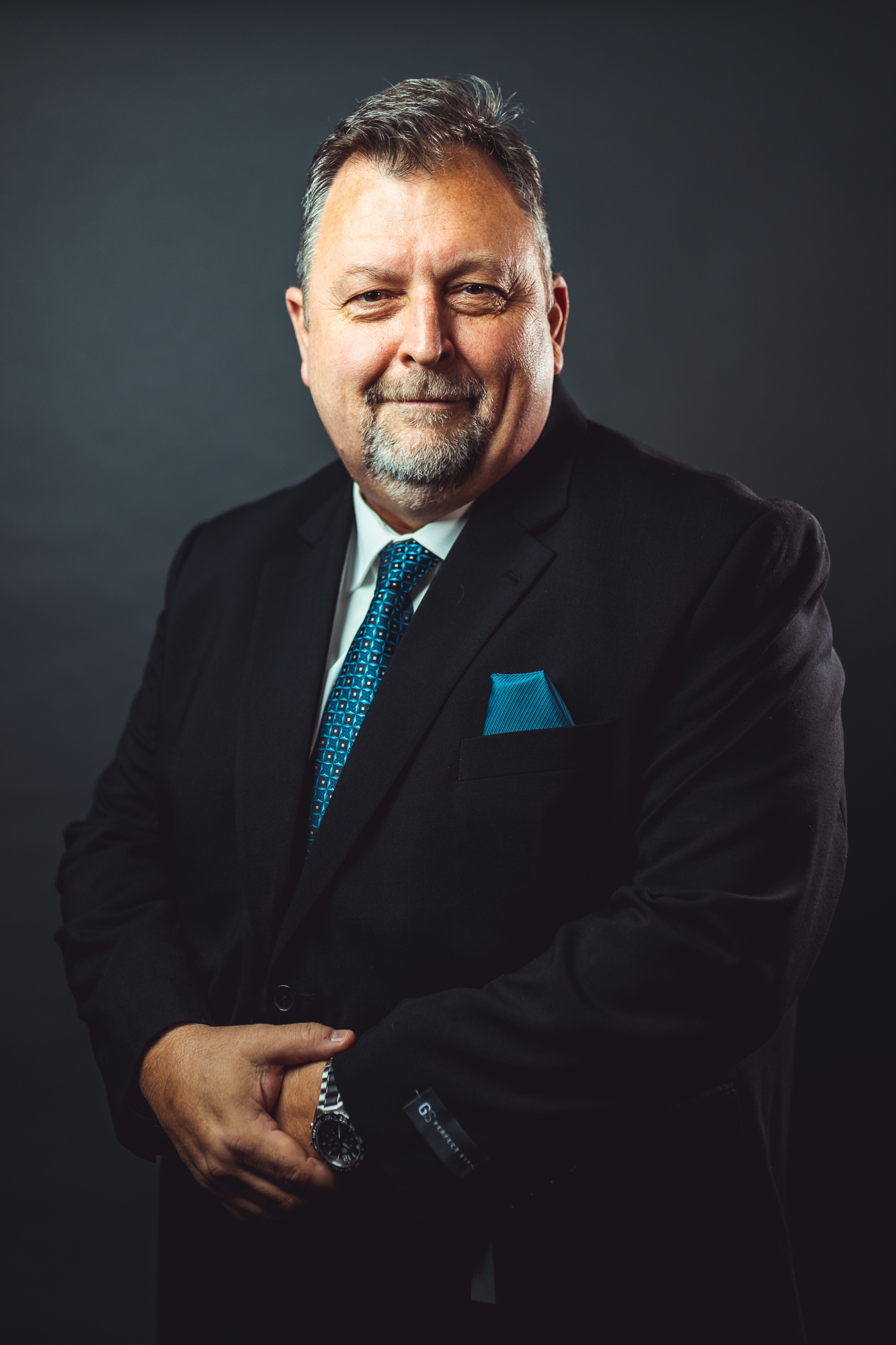 Professional headshot of Gary Bland, Vice President at GRP|WEGMAN, smiling with a black suit jacket, white shirt and a blue tie and pocket square, in front of a neutral gray background