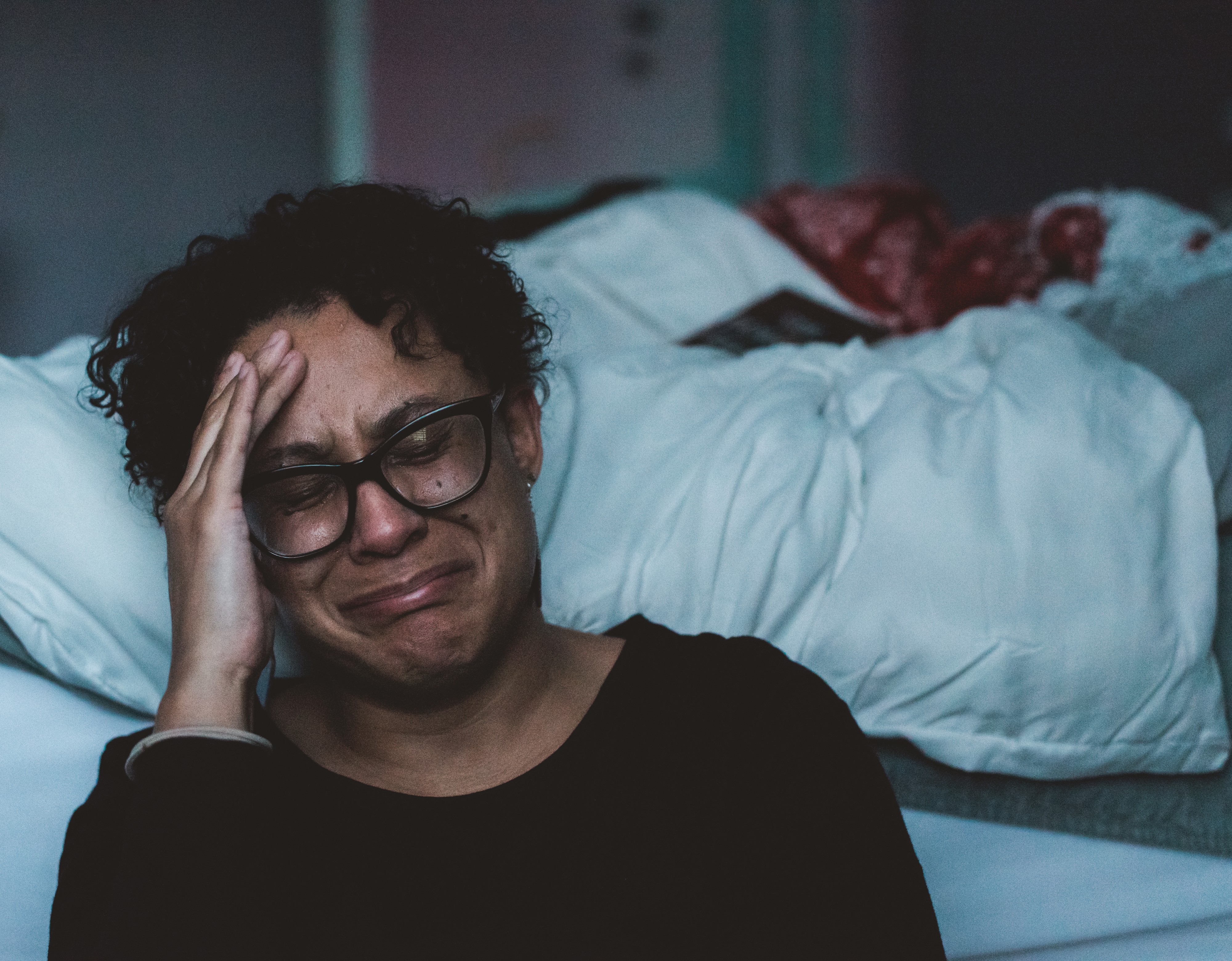 A person looking like they’re about to cry, holding their hand to their head, sitting in front of their bed