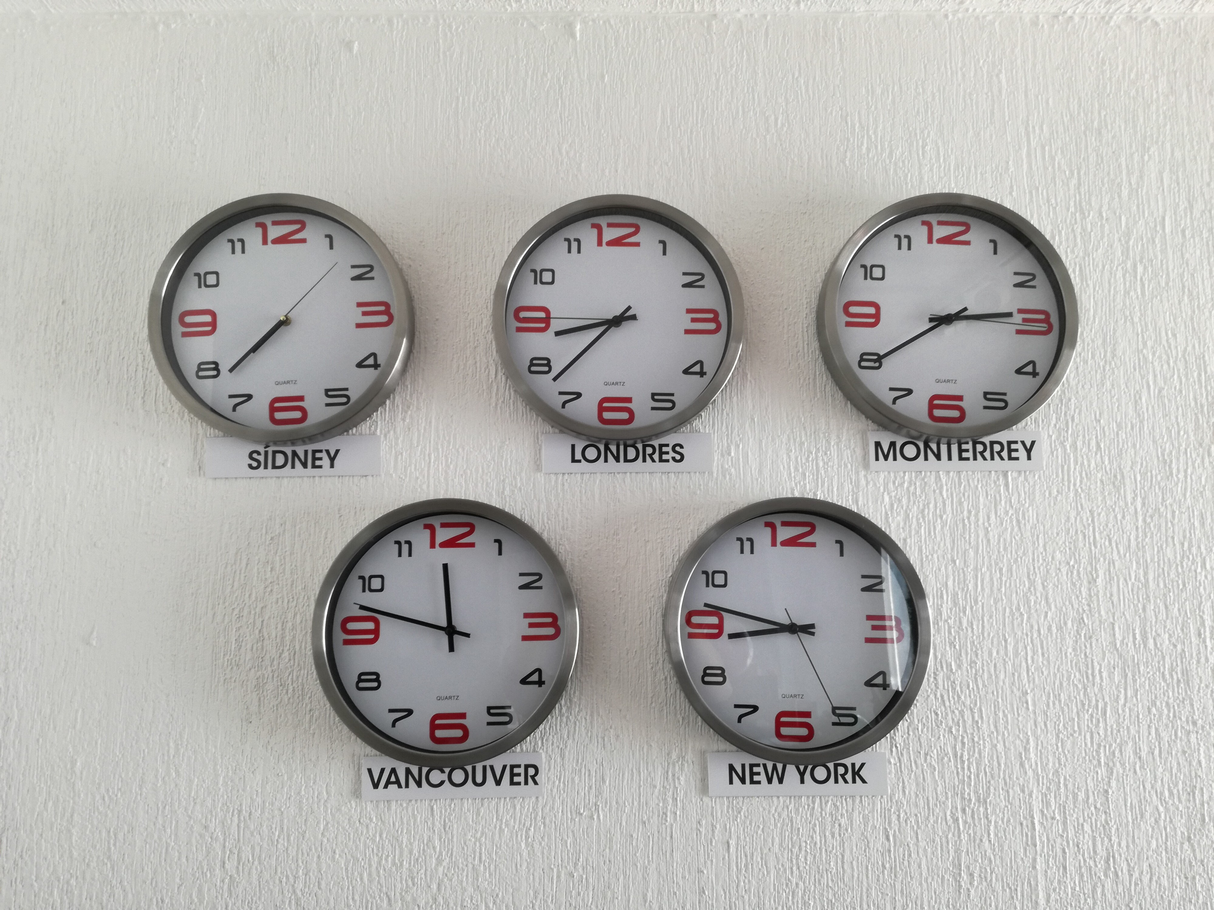 Five clocks hanging on a wall, pointing at different time zones.