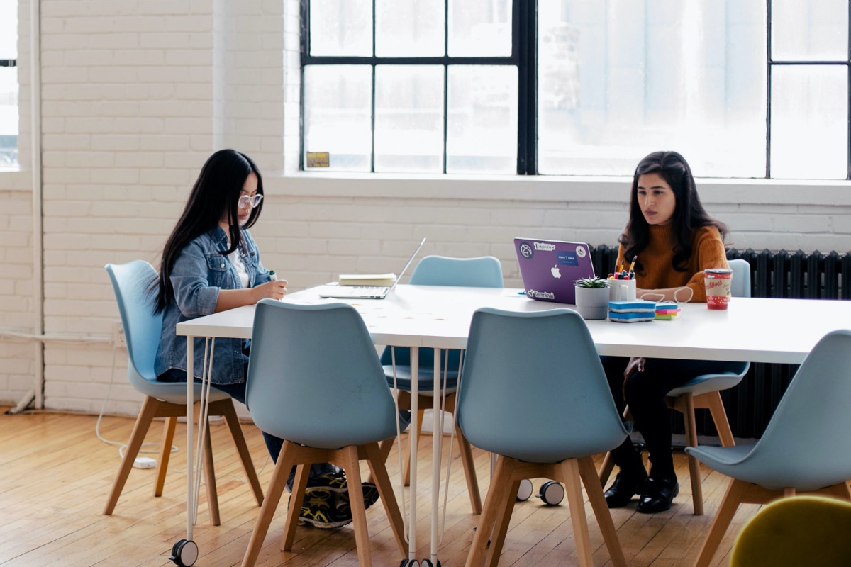 Two females working at a communal table with laptops.