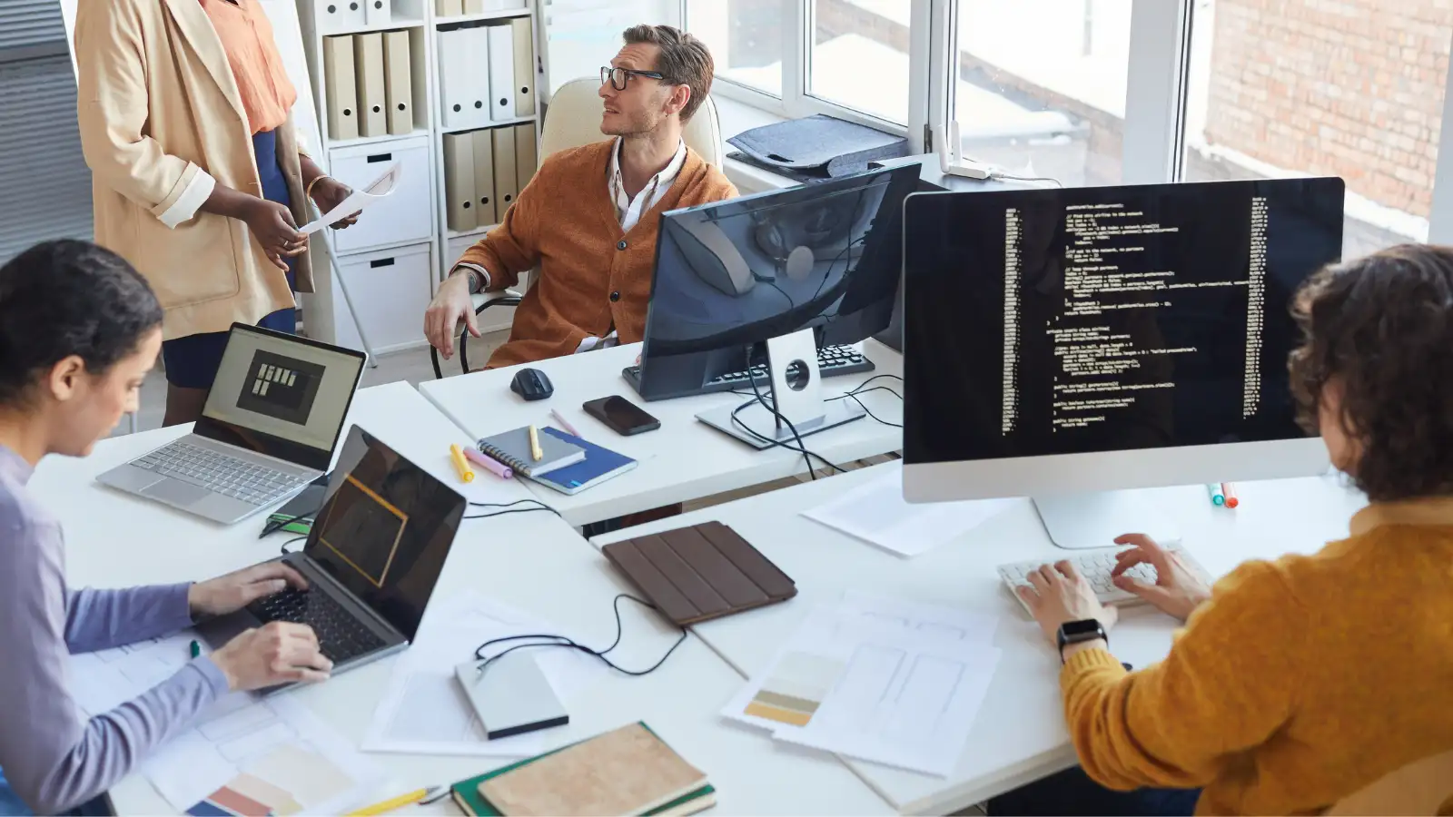 Group of young full stack developers in a casual office setting typing coding languages into computers and discussing projects.