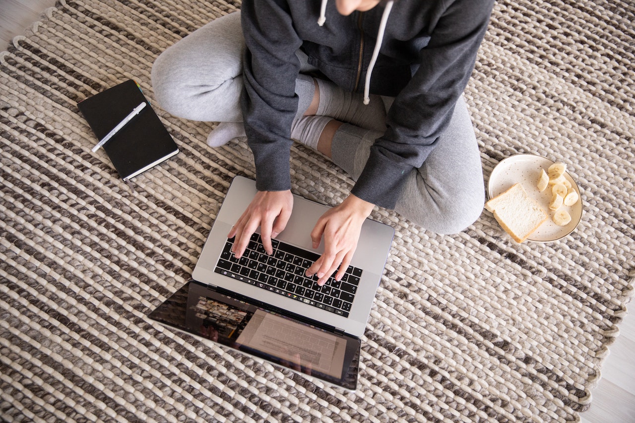 An overhead shot of a person working on a laptop. They're sitting on the floor, on top of a lovely monochrome rug. Beside the person is a black notebook with a pen, and suspiciously, a plate of bread with sliced bananas.