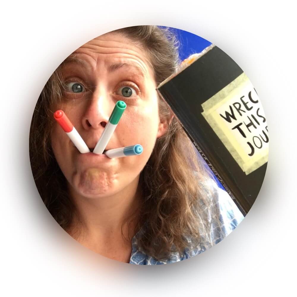 Woman with pens in her mouth holding a journal titled "WRECK THIS JOURNAL"