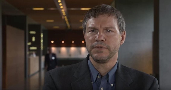 Homem branco usando terno preto e camisa azul com a recepção de um prédio ao fundo