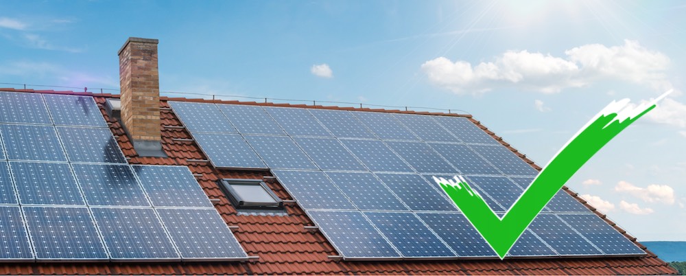 Eine PV-Anlage auf rotem Hausdach, mit blauem Himmeln und einem grünen Haken im Vordergrund