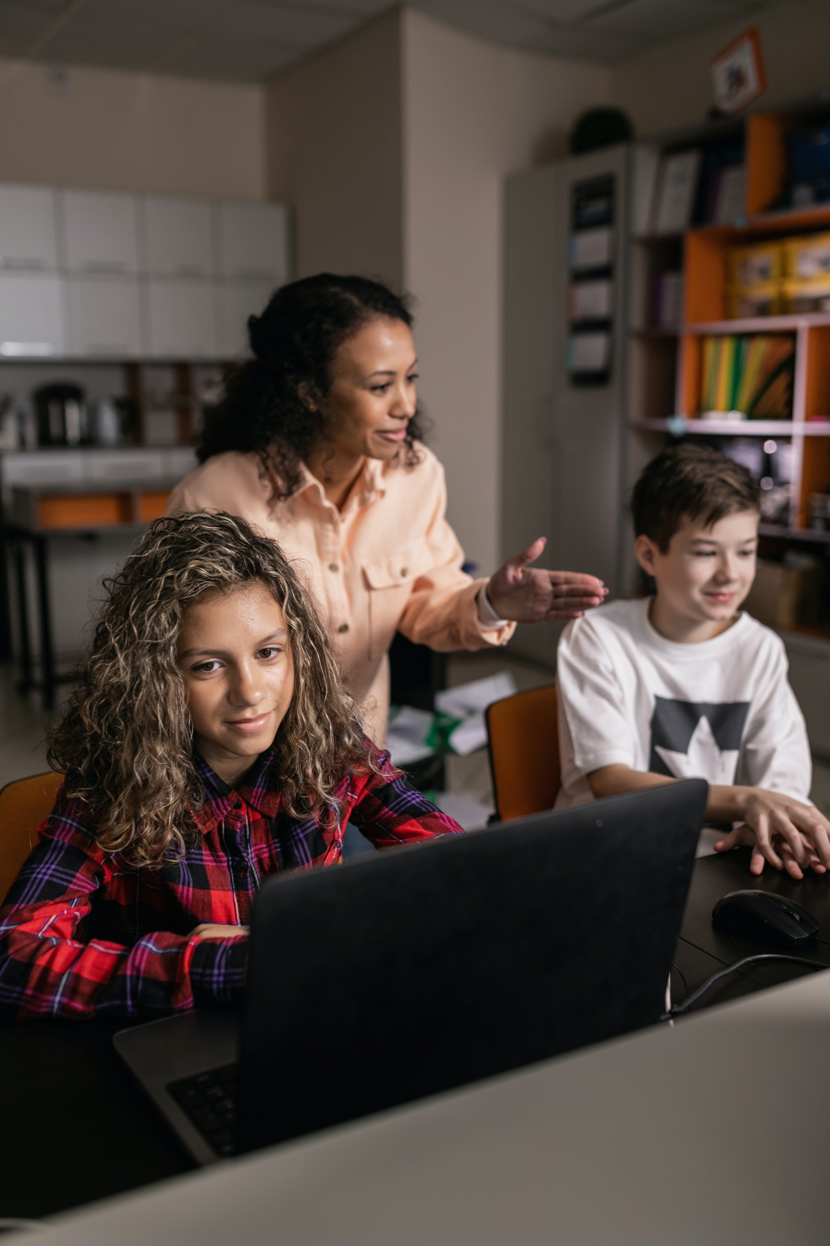 A teacher helping 2 students who are doing class work on the computer. One student is a girl wearing a plaid button up shirt. The other is a boy wearing a white 2 shirt.