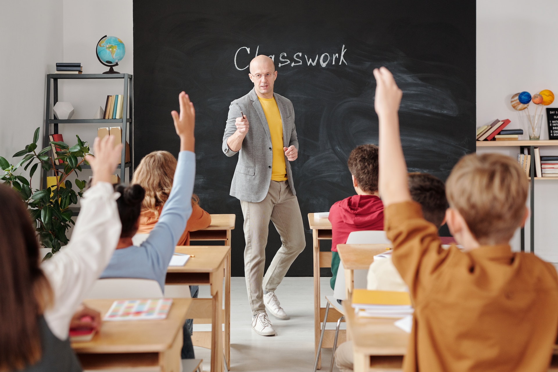 Classroom of kids raising their hands while a male teacher at the front of the classrooms point to a student to hear their answer. 