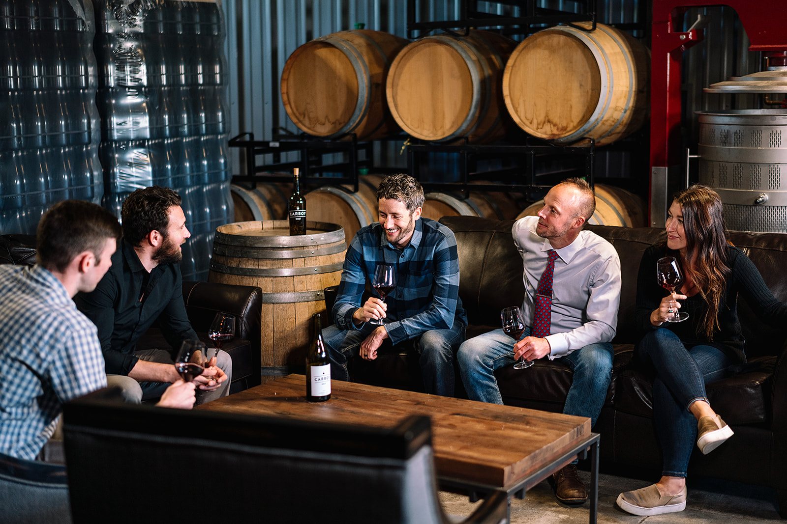 Carboy Winery staff sits on a couch and chairs and talks while drinking a glass of wine in front of kegged wine.