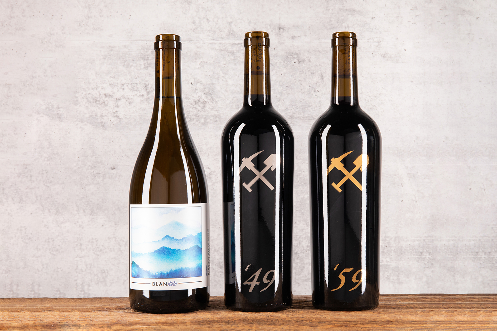 Three bottles of Carboy Winery wines sit on a wooden table with a concrete background. 