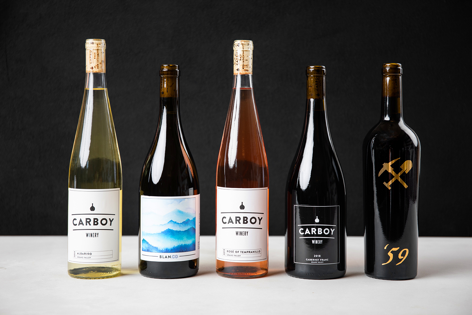 Five bottles of assorted Carboy Winery wines sit on a marble counter with a black background. 