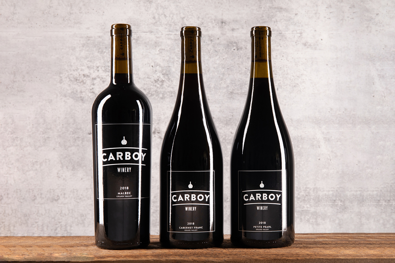 Three bottles of Carboy Winery wines sit on a wooden table with a concrete background. 