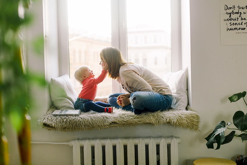 Mum and baby sitting in a window seat, the baby is cupping its mum's face.